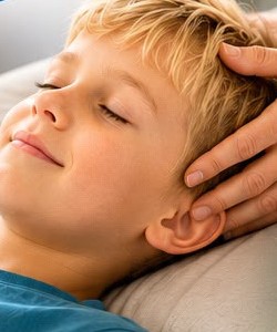 A child lying back with eyes closed while hands rest lightly on his head